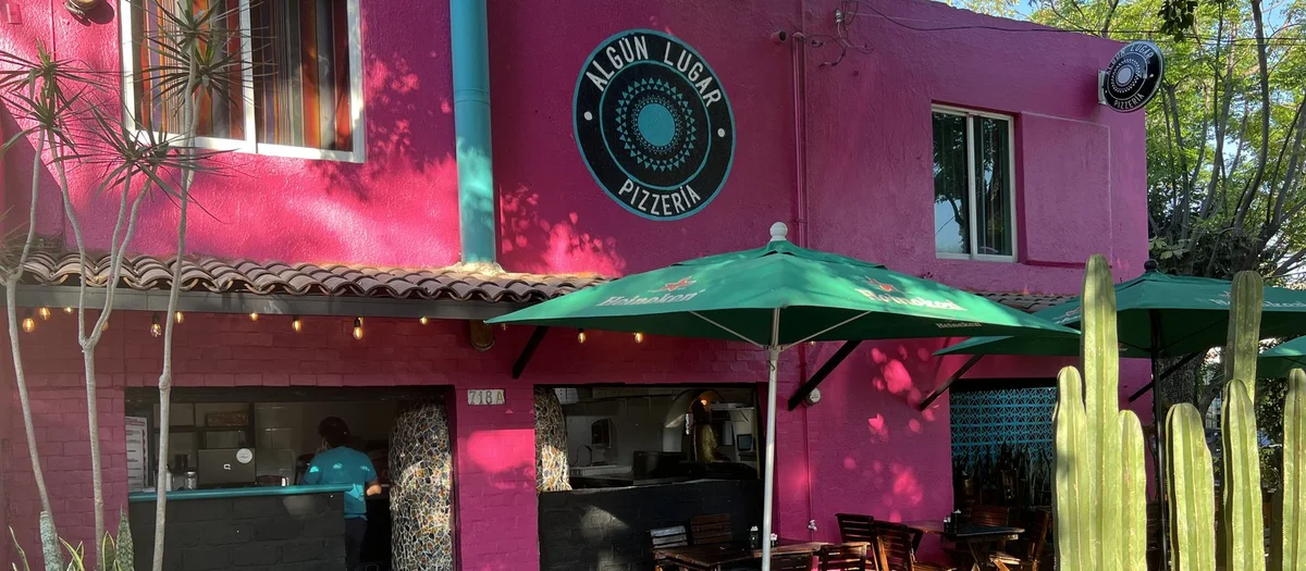 Fachada rosa de Algún Lugar Pizzería con letreros circulares, sombrillas verdes y cactus.